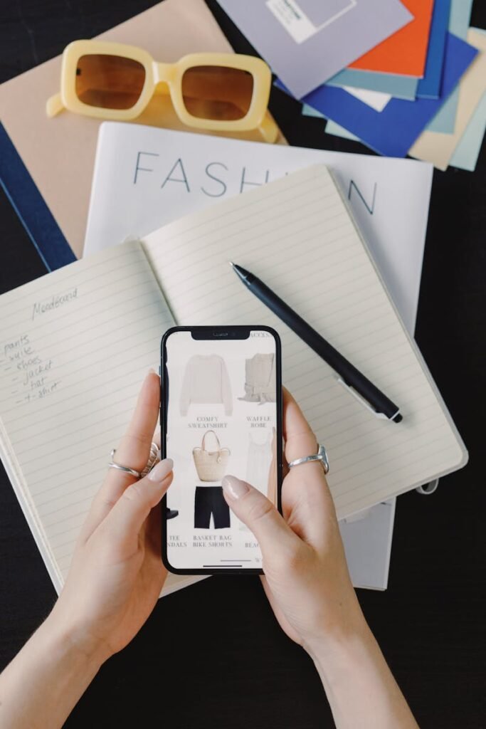 Person using smartphone for online fashion shopping at a desk with notepad and glasses.